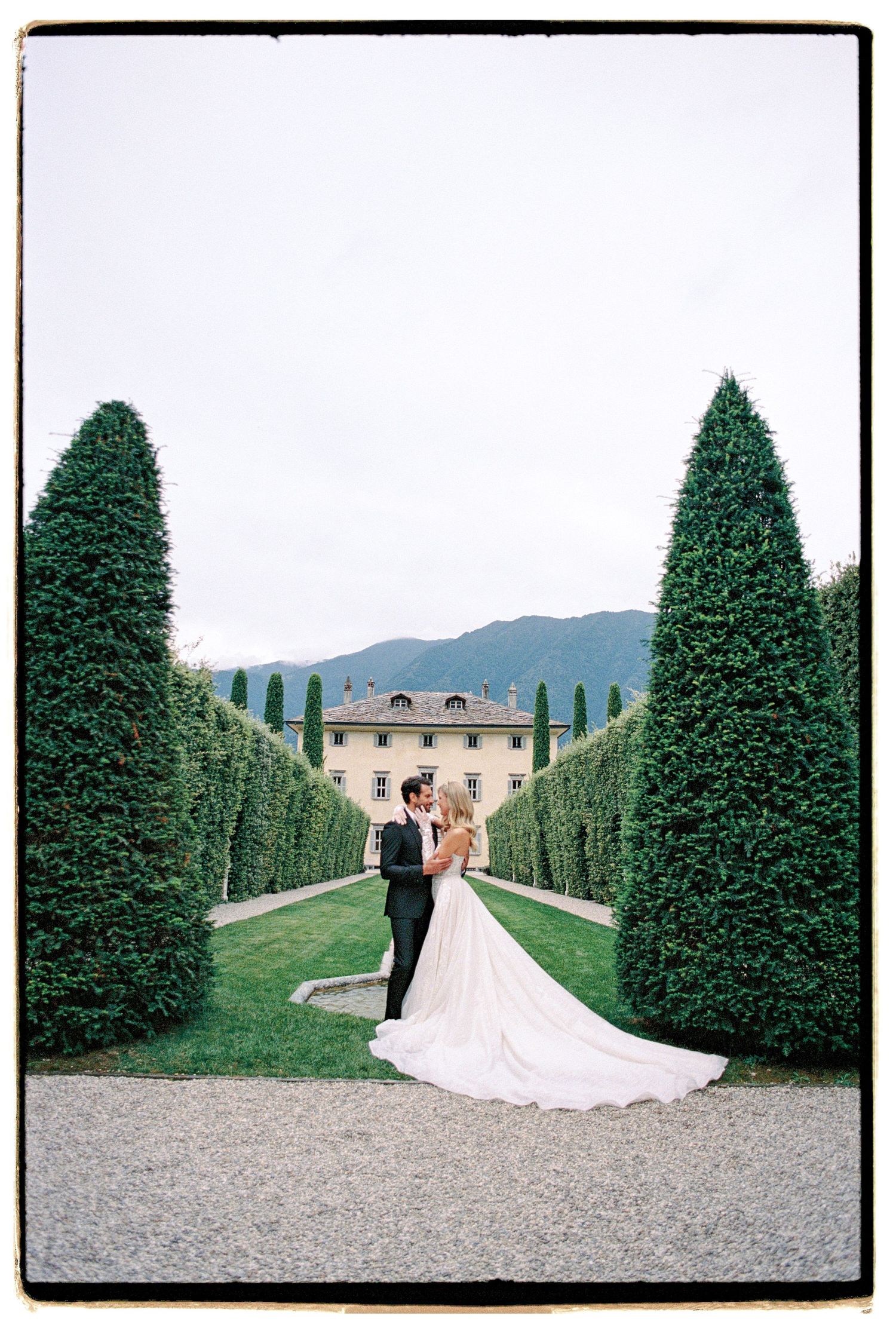 Manicured garden path lined with tall cypress trees leads to a grand villa in the mountains.