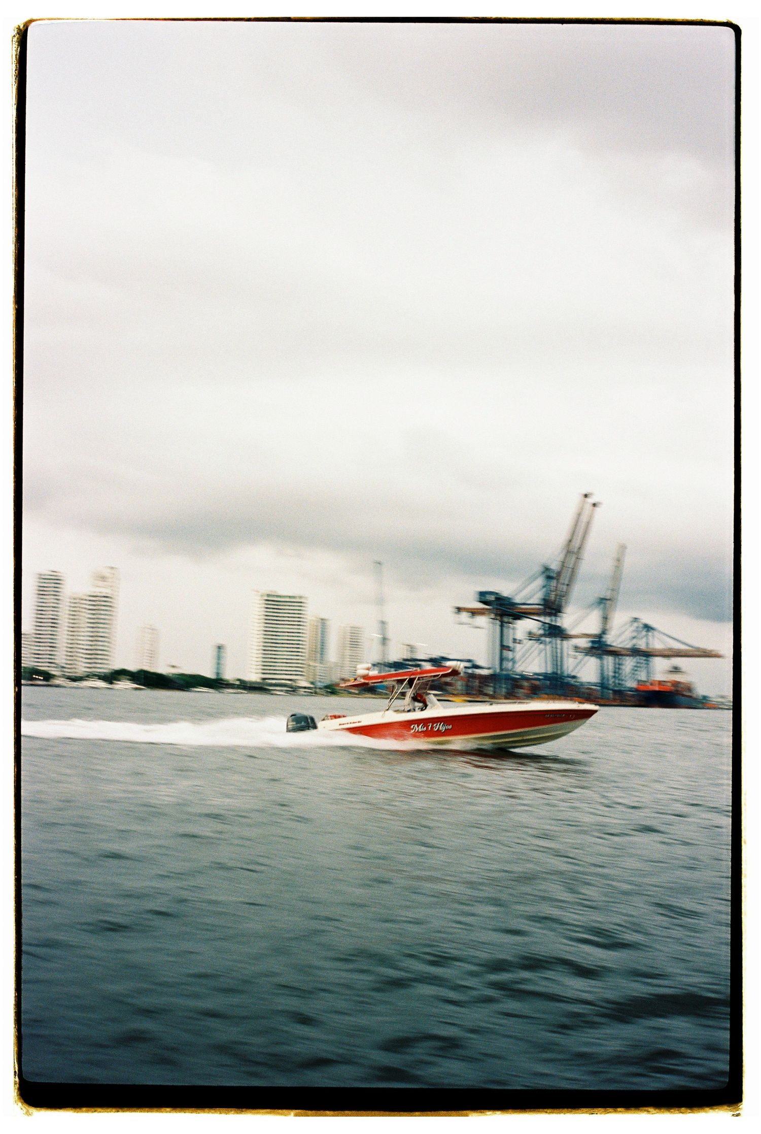A red speedboat racing across blue waters with industrial port cranes in the background.