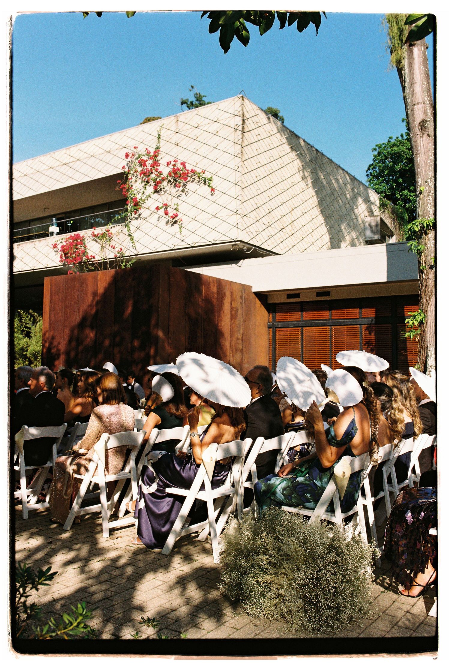 Wedding guests seated in white chairs wearing matching white sunhats at an outdoor ceremony.
