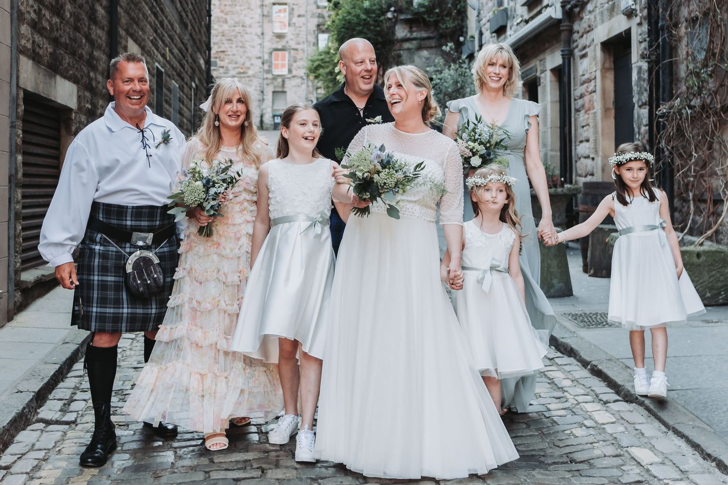 Wedding party in Scottish attire posing on a historic cobblestone street.