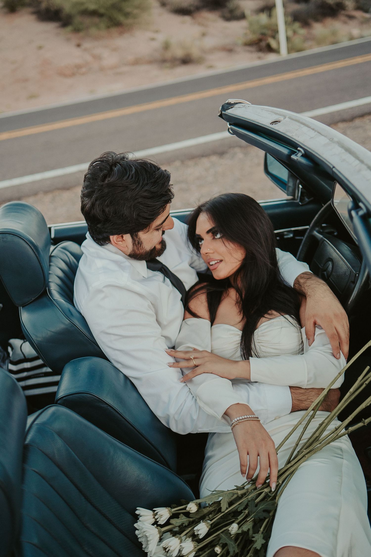 Newlyweds embracing in vintage convertible against desert landscape.