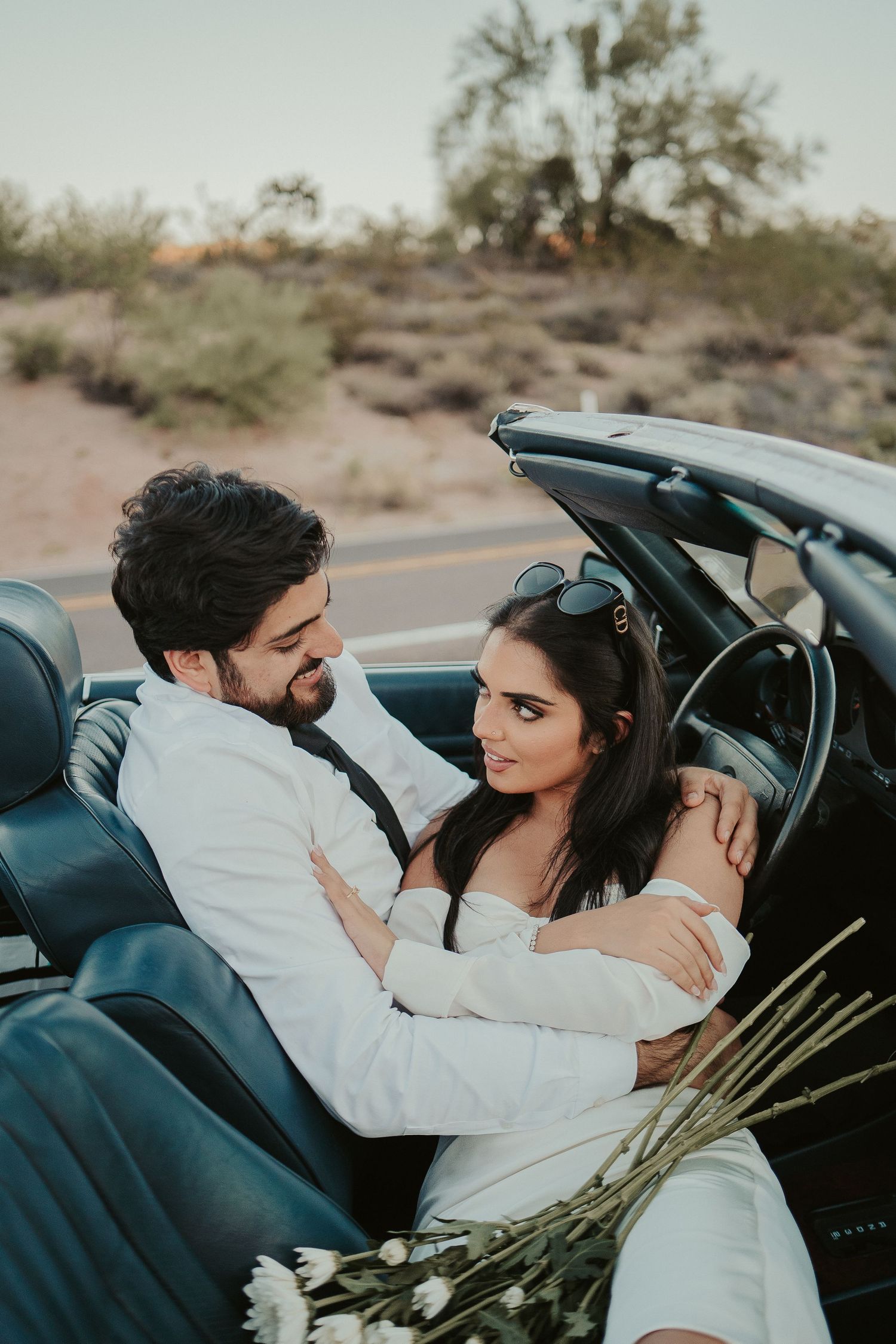 A couple embraces in the front seat of a convertible against a desert landscape backdrop.