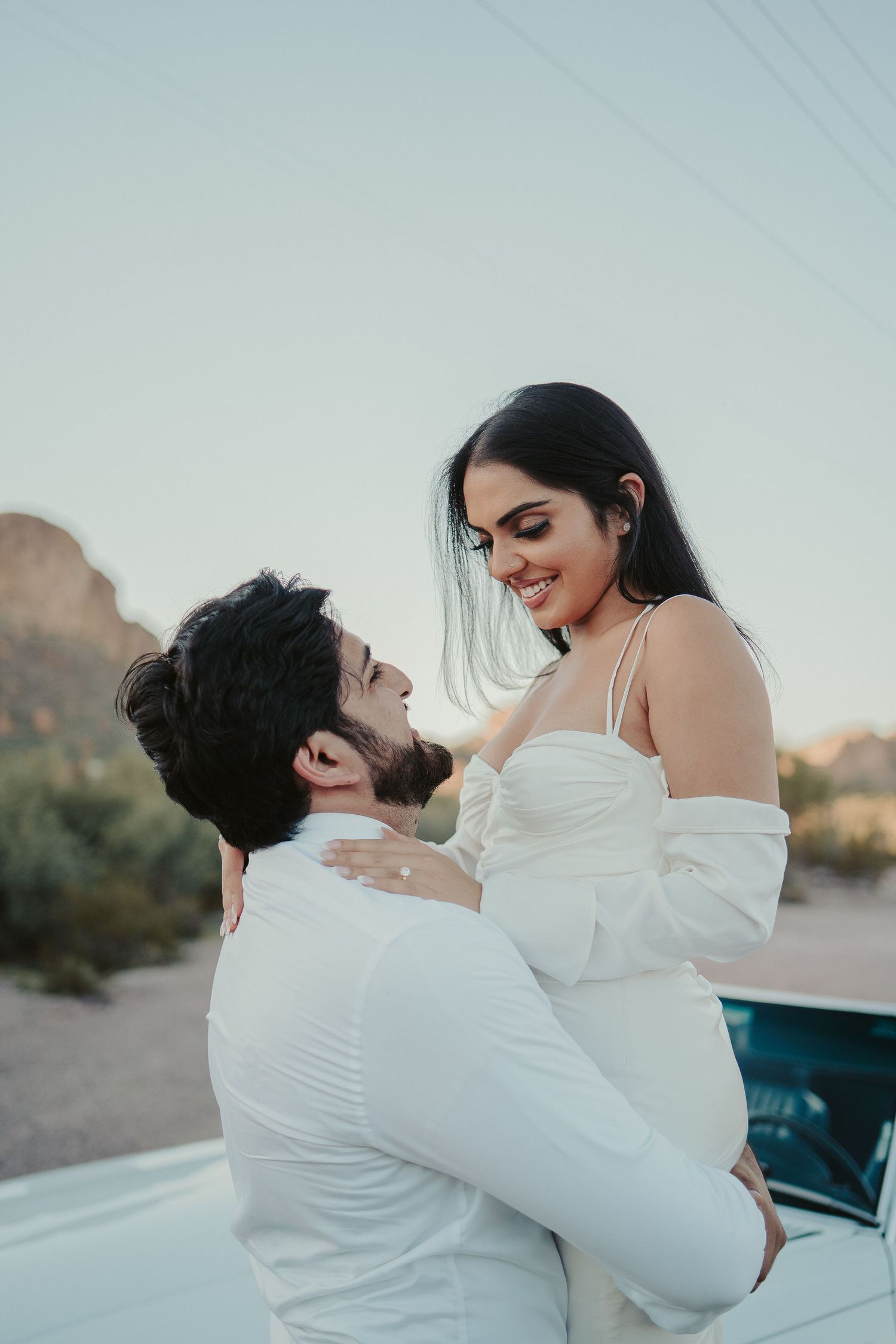 Couple in formal wear shares playful moments near a classic white car in desert setting.