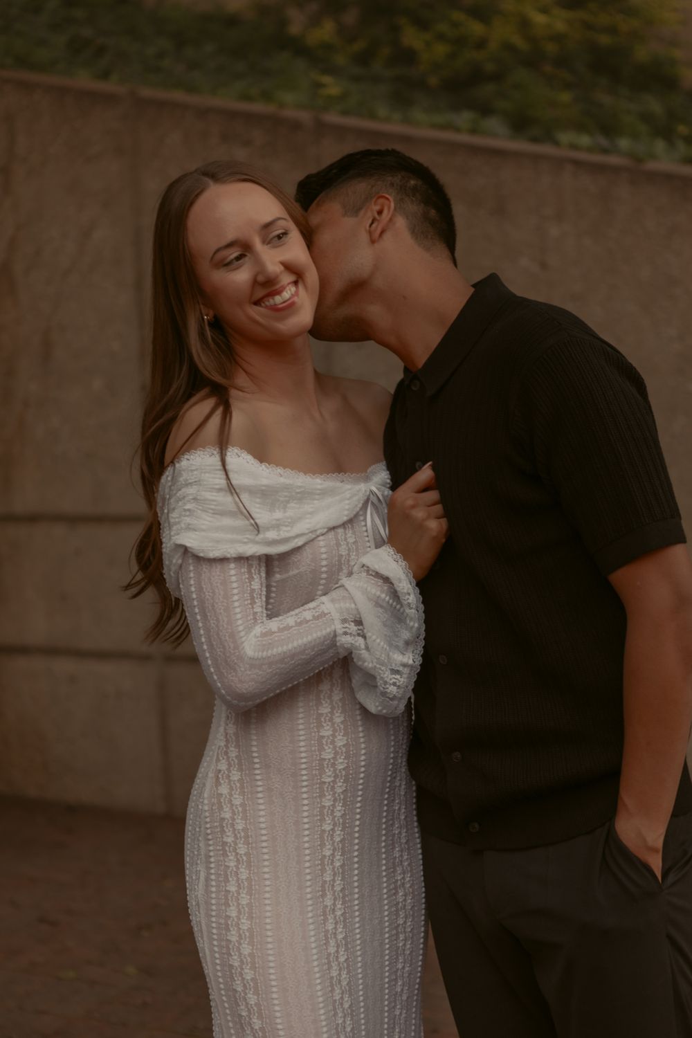 A romantic couple embraces outdoors at sunset, with one person wearing a white knit sweater dress and the other in black.