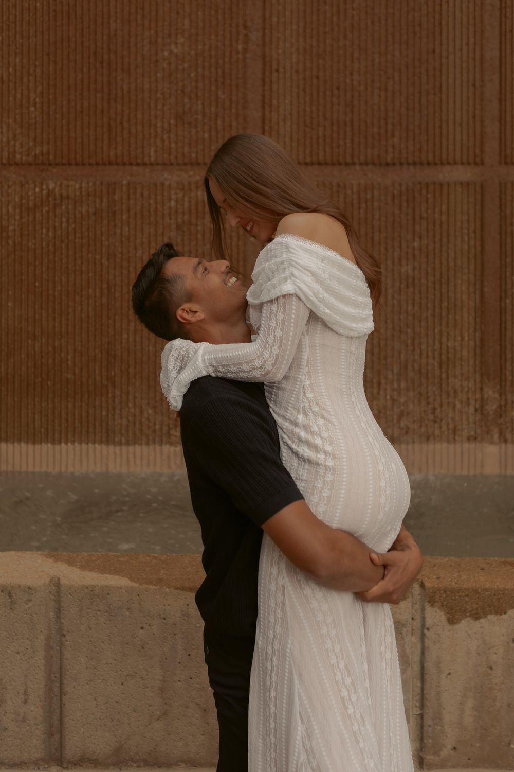 A romantic couple shares an intimate moment as he lifts her in a white dress against a textured wall background.