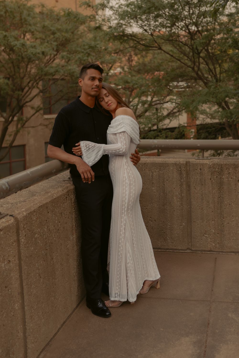A romantic couple embraces on a concrete balcony at sunset, with one person wearing a flowing white dress.