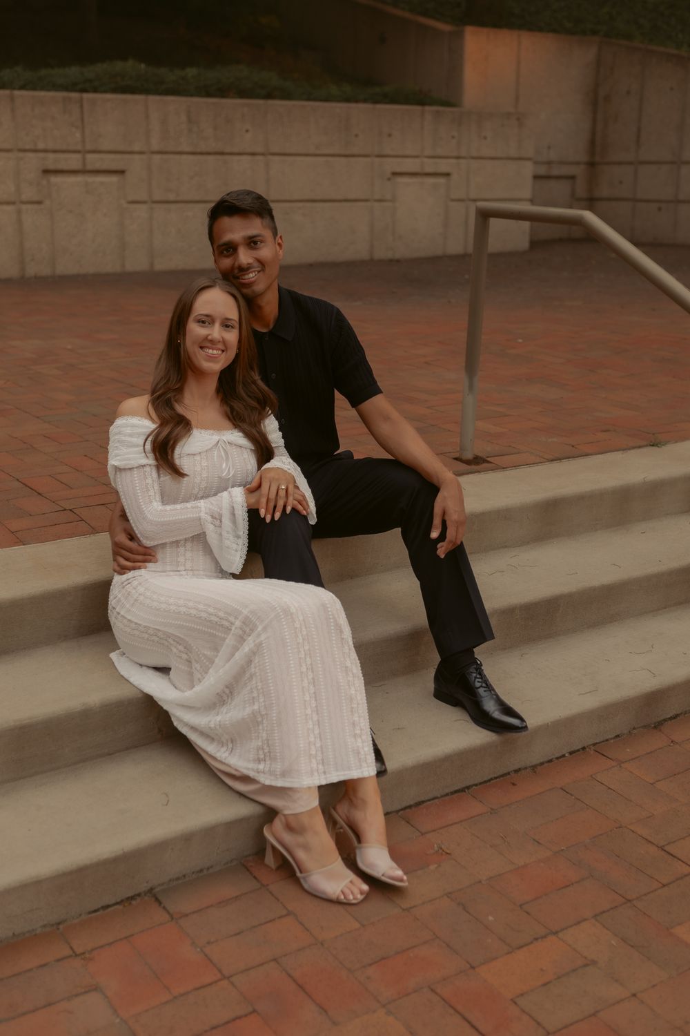 A romantic couple sits on brick steps, with the woman wearing a white dress and man in black casual attire.