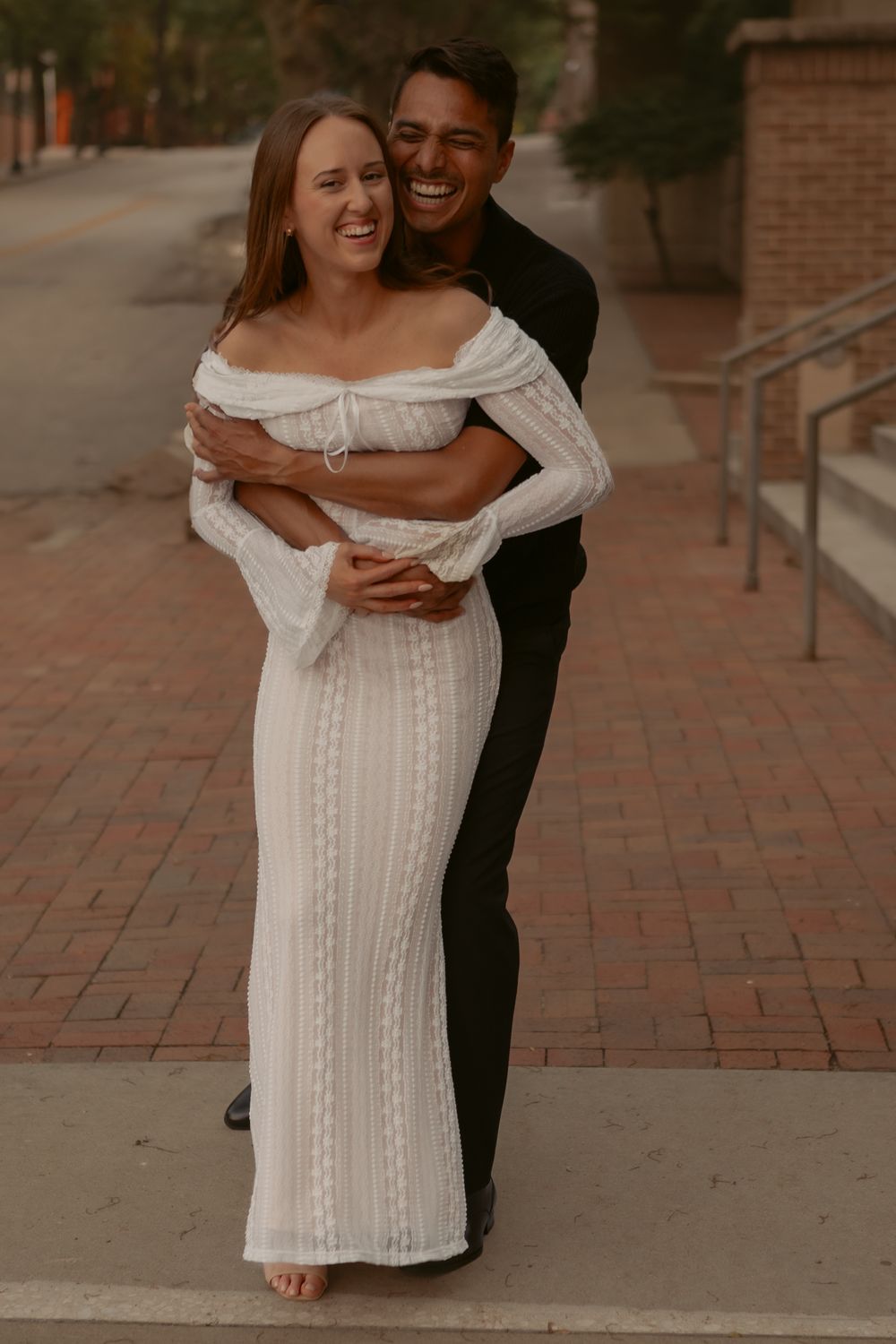 Romantic couple embraces on brick walkway with woman in white off-shoulder dress and man in black outfit.