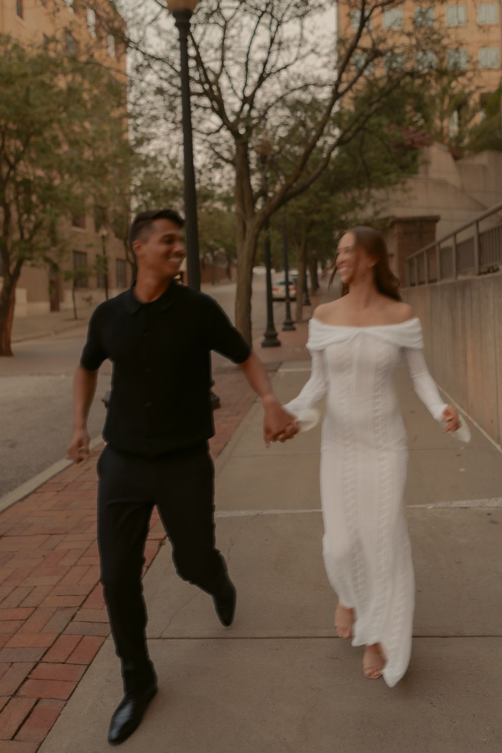 Playful couple runs and dances along a brick pathway with trees and buildings in background.