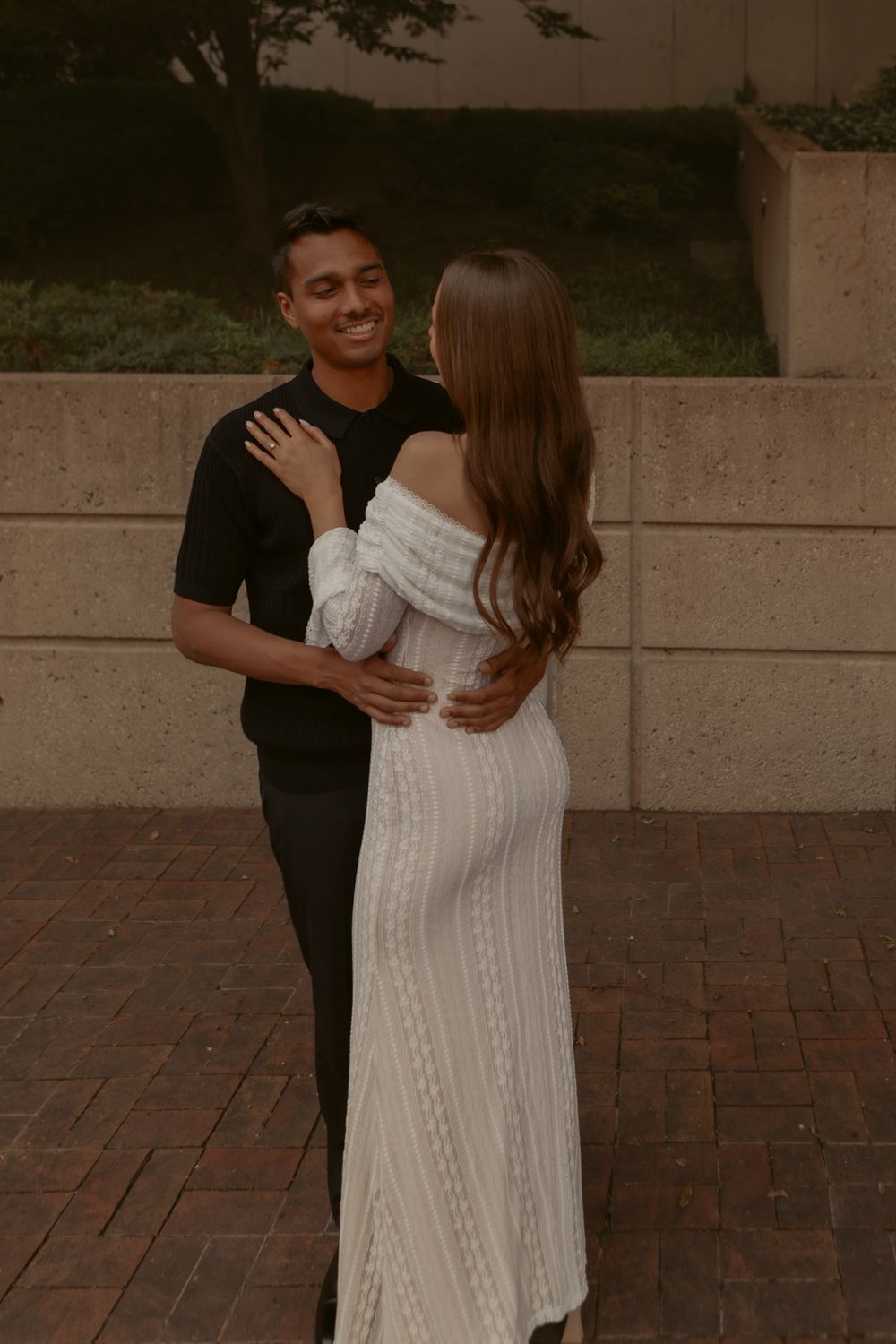 A couple embraces on a brick pathway, with the person wearing an elegant white off-shoulder dress standing close together.