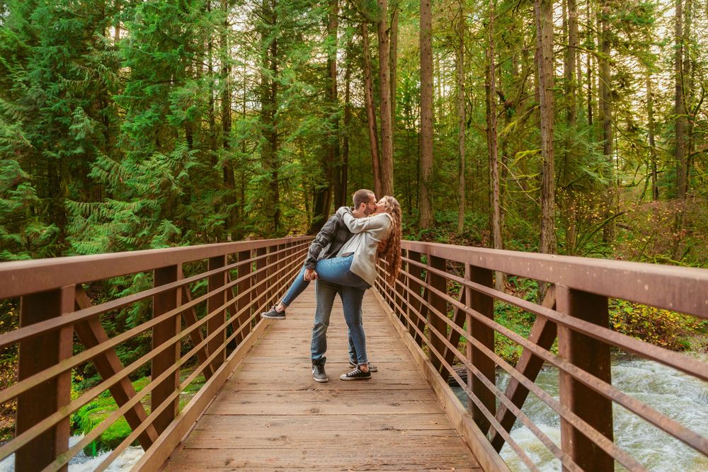 Young couple sharing a romantic moment on a wooden footbridge surrounded by towering evergreen trees in nature.