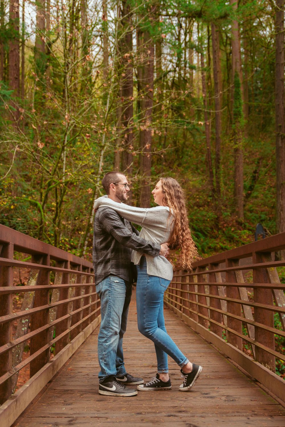 Couple embraces romantically on wooden footbridge surrounded by lush evergreen forest in Pacific Northwest.
