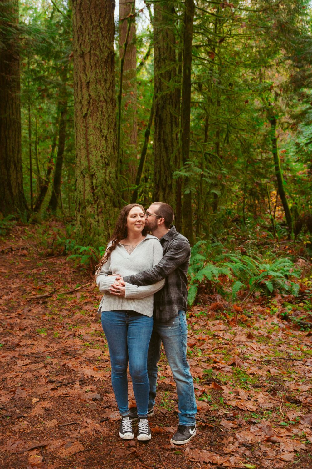 A couple embraces while standing on a forest trail surrounded by tall evergreen trees and fallen autumn leaves.