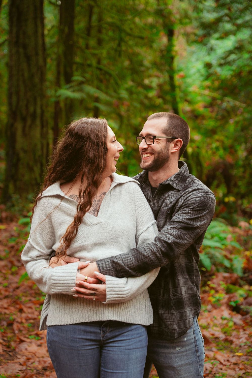 A couple dressed casually embraces and laughs together in a lush forest setting during autumn.
