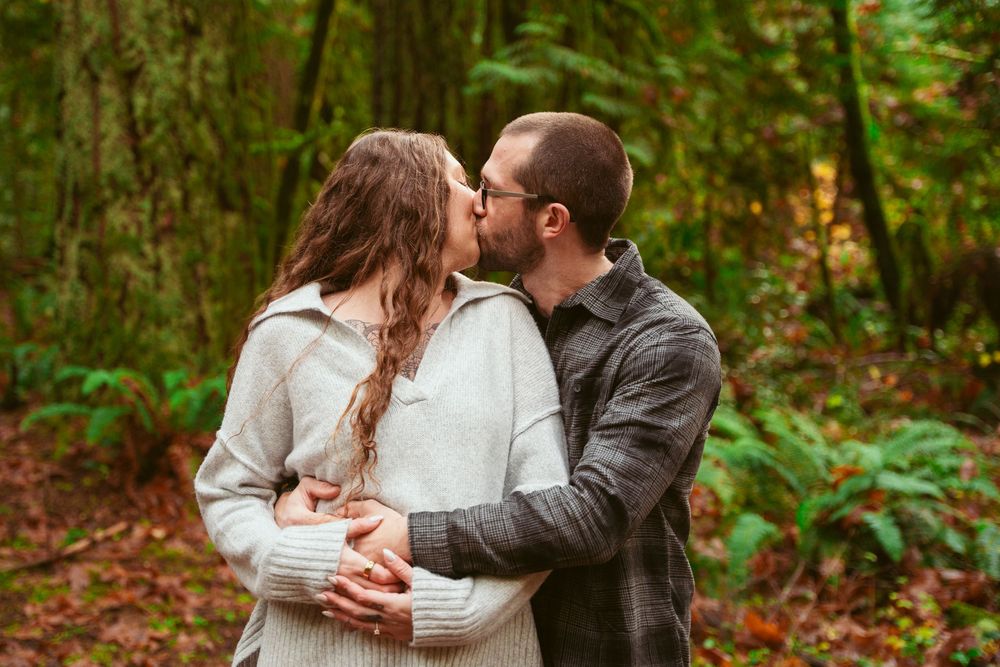 A couple in warm sweaters embrace while sharing a tender moment in an autumn forest setting with lush greenery.