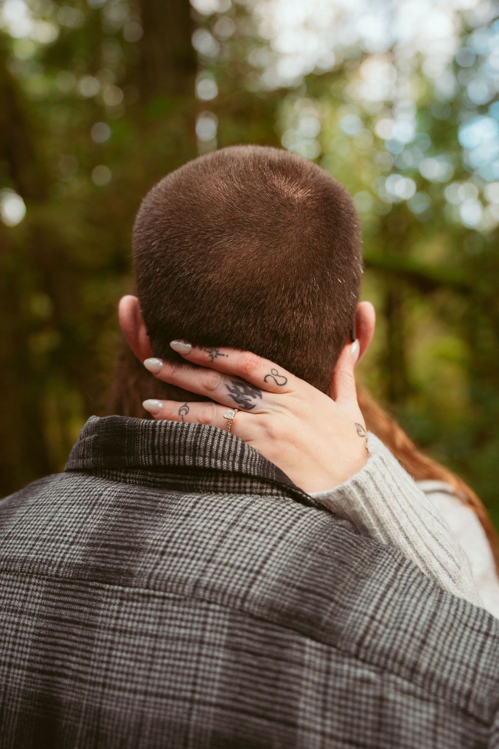 Hand with an engagement ring reaches around to touch the back of a neck over a plaid suit jacket in an outdoor setting.