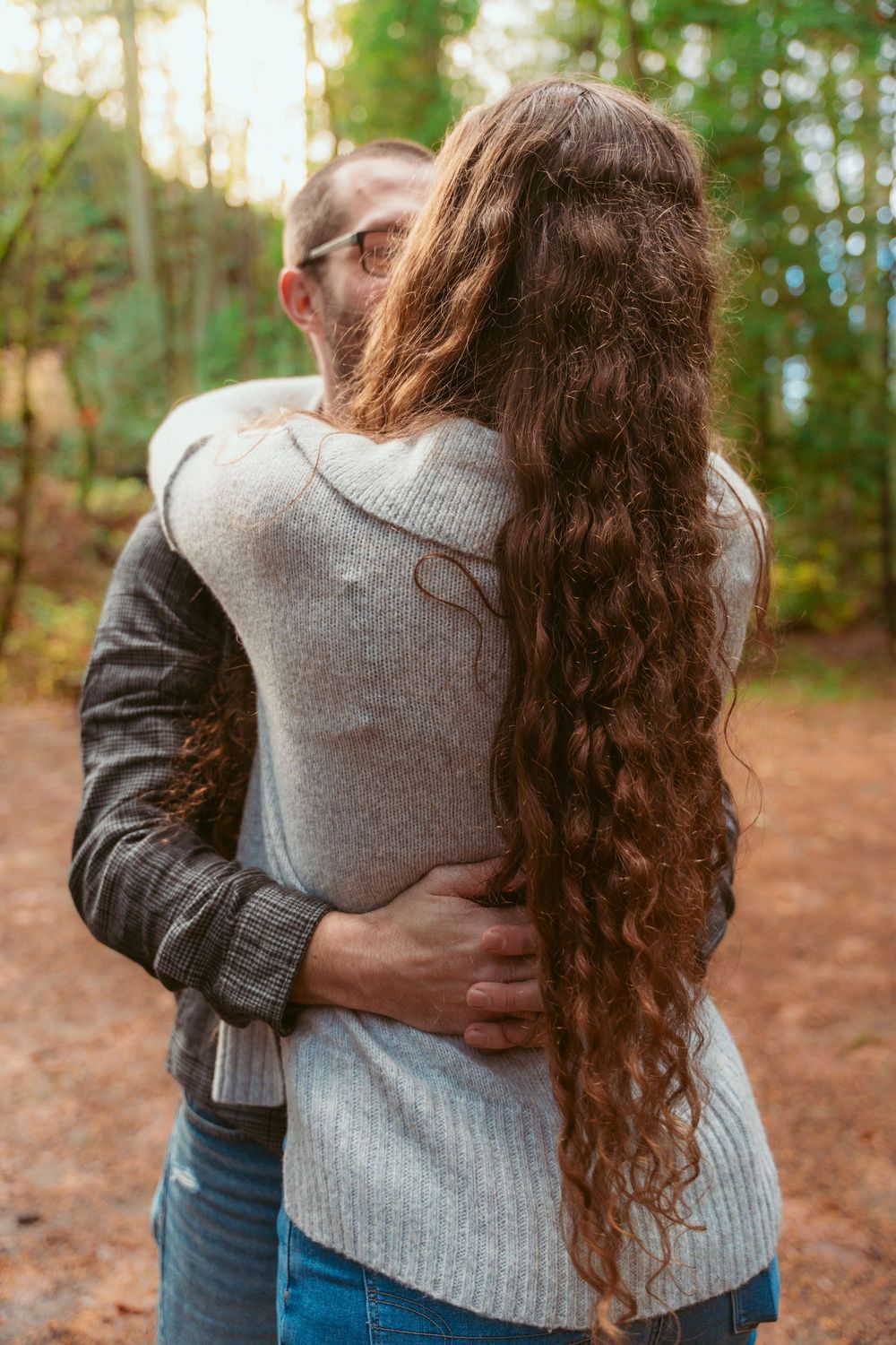 A couple embraces on a forest trail, with one wearing a gray sweater and the other in a leather jacket.