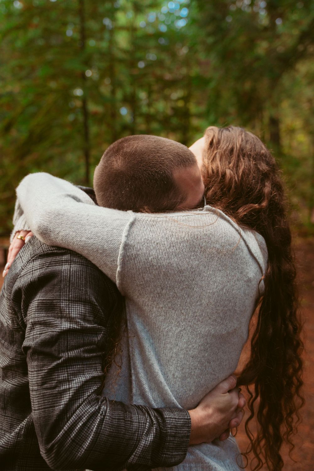Two people in intimate embrace share a romantic moment in a wooded forest setting wearing casual sweaters and coats.