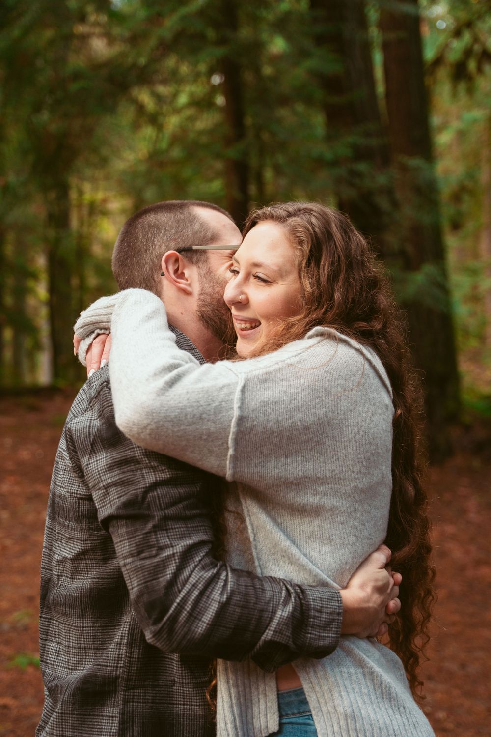 A couple shares an intimate embrace while laughing together on a wooded trail surrounded by trees.