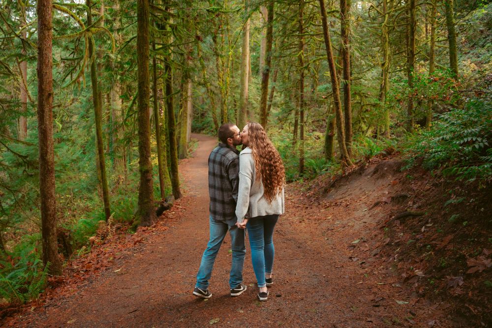 Two people stand together on a winding forest trail surrounded by tall evergreen trees on an autumn day.