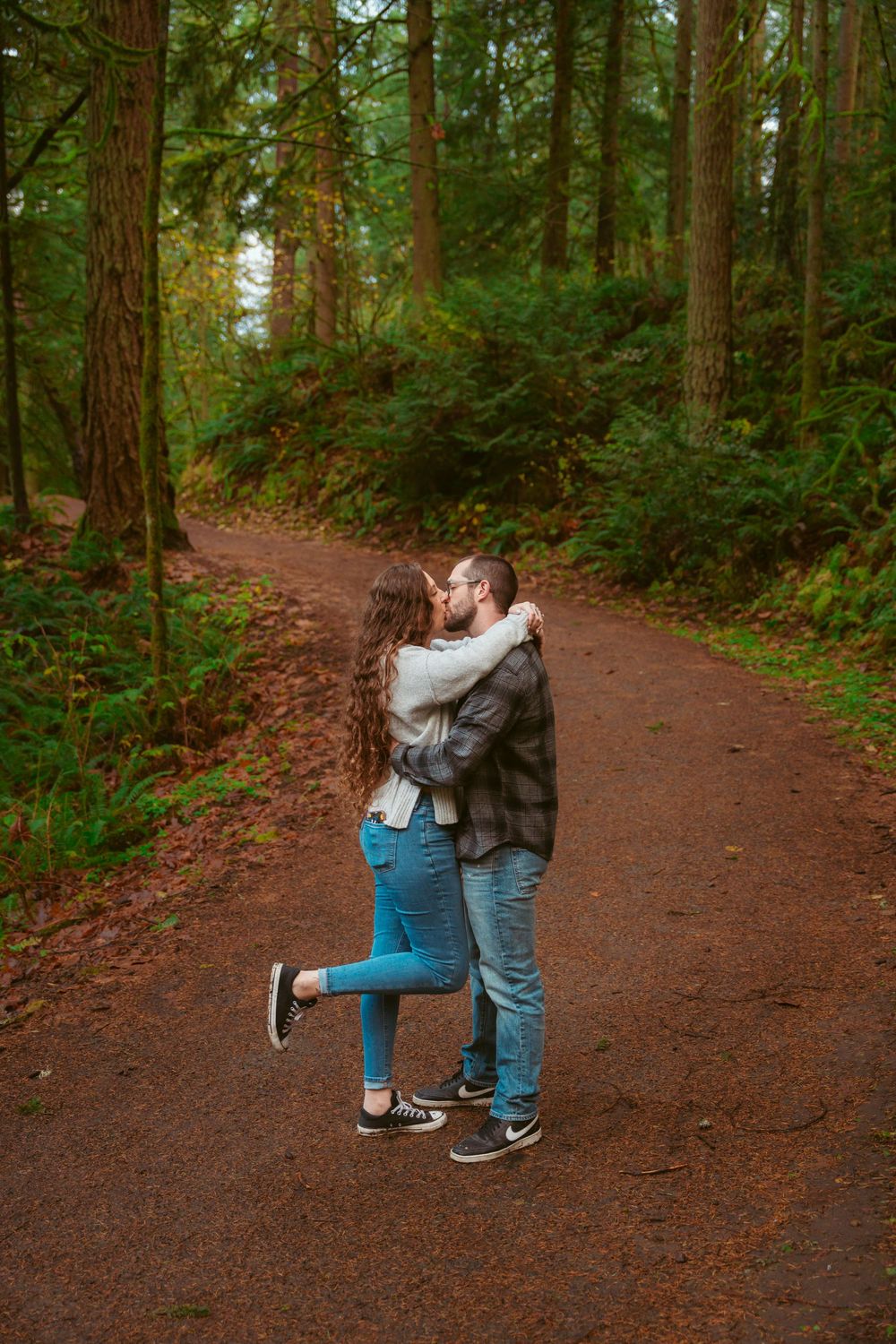 A couple shares a romantic embrace while standing on a dirt trail through a lush evergreen forest.