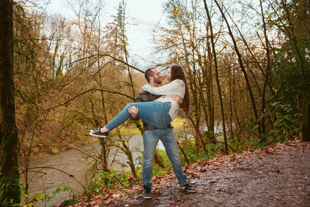 Two people embrace joyfully on a forested hiking trail surrounded by autumn trees and fallen leaves.