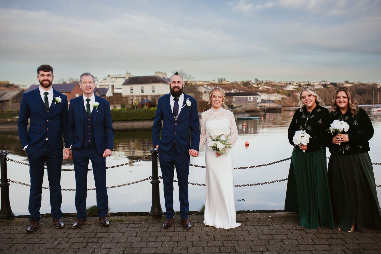 Wedding party in navy and dark green attire poses on rooftop deck overlooking city buildings during sunset.