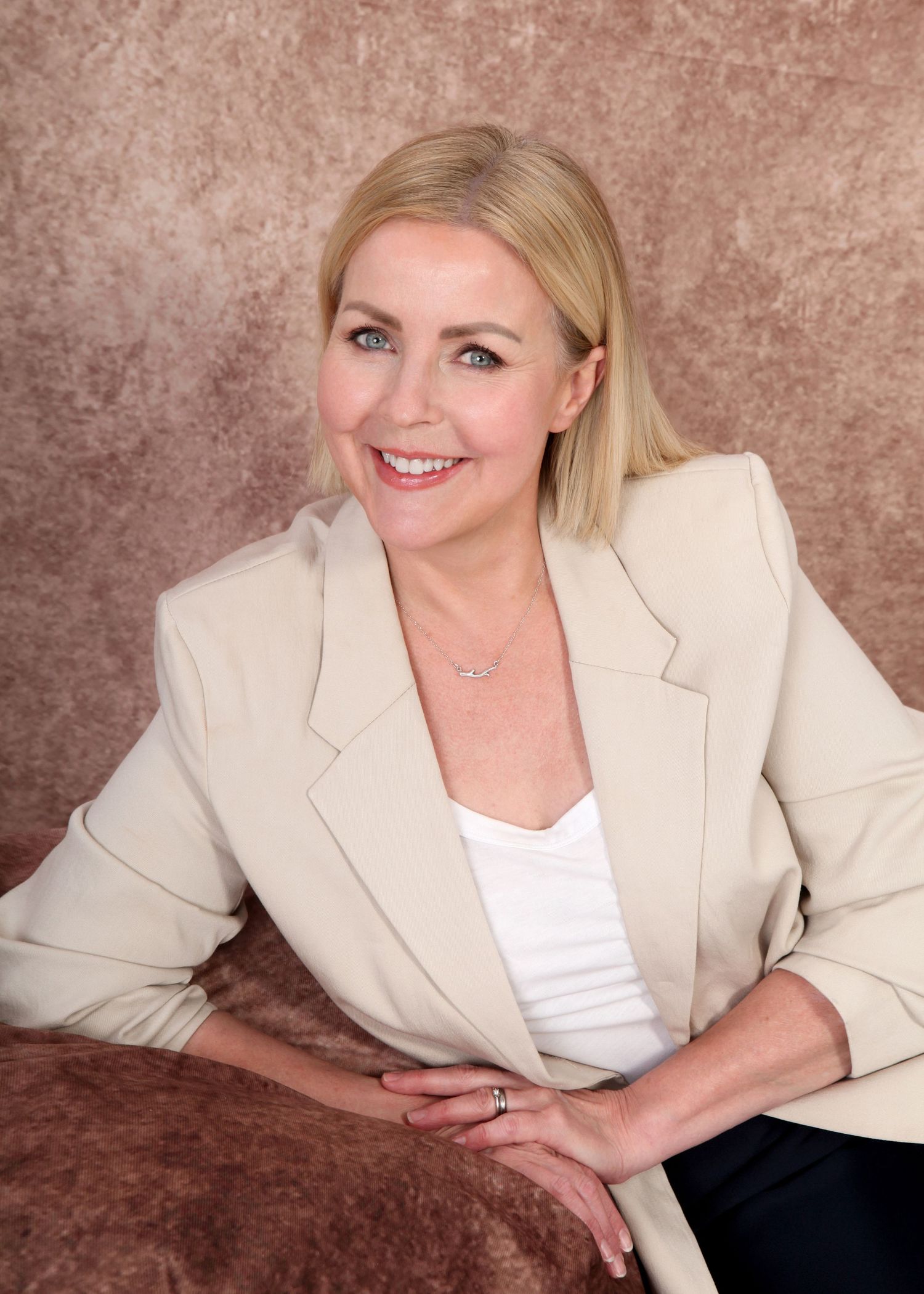 Professional portrait of someone in a beige blazer and white top posing against a brown textured backdrop.