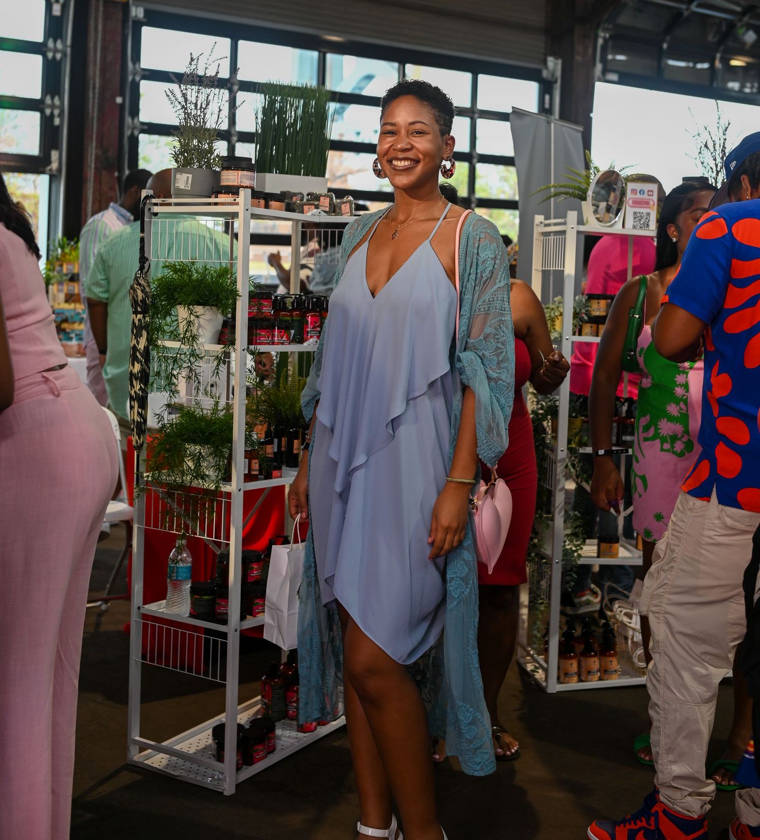 Smiling person in flowing gray dress stands in retail space surrounded by colorful displays and shelving units.