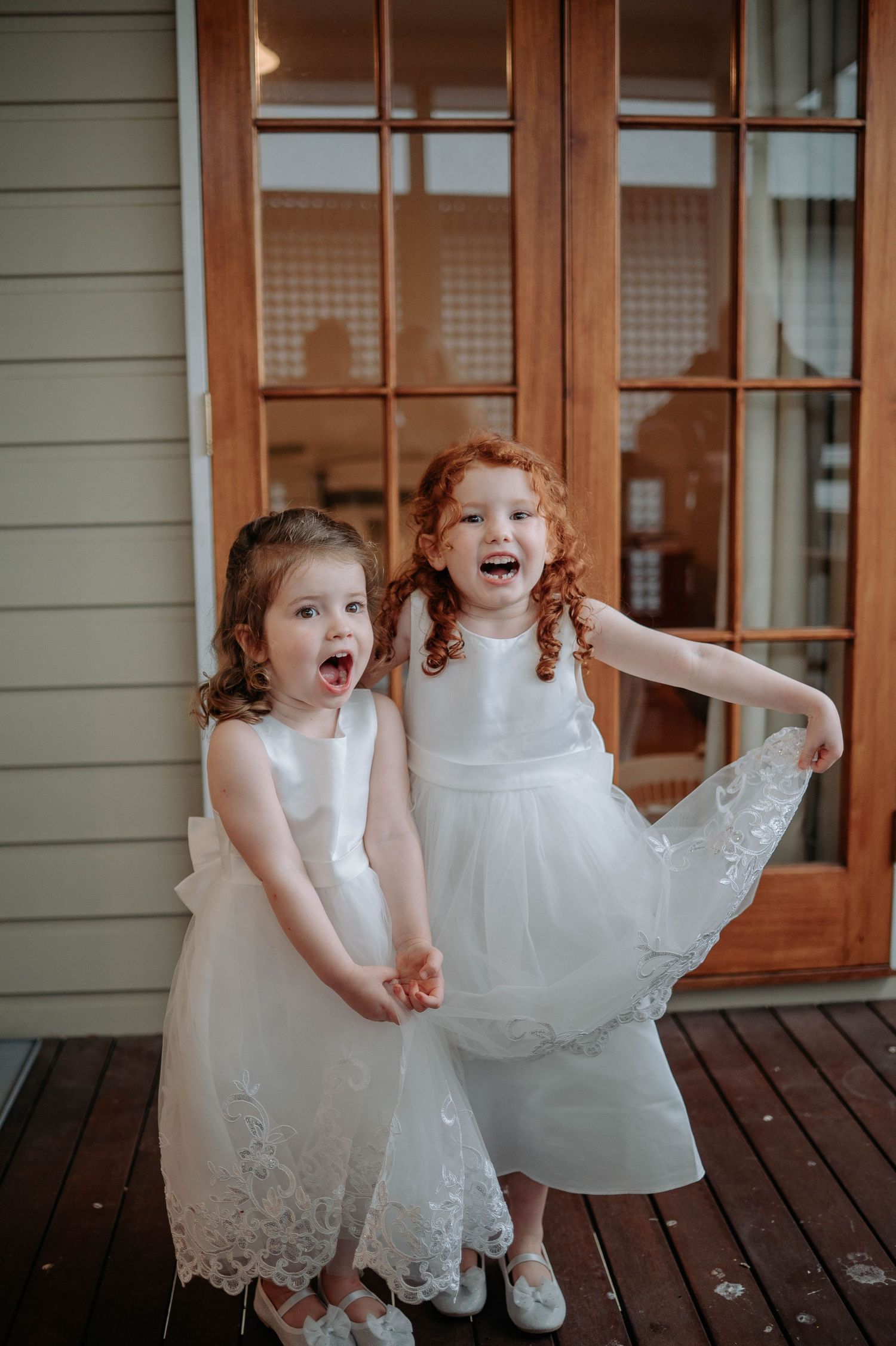 Two young flower girls in white lace dresses laugh excitedly while standing on a wooden deck outside a house.