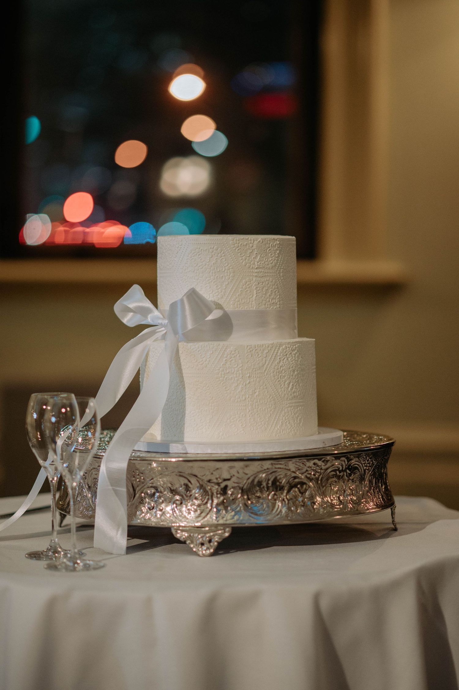 Two-tiered white wedding cake with delicate embossing sits on ornate silver stand with white ribbon bow against blurred lights.