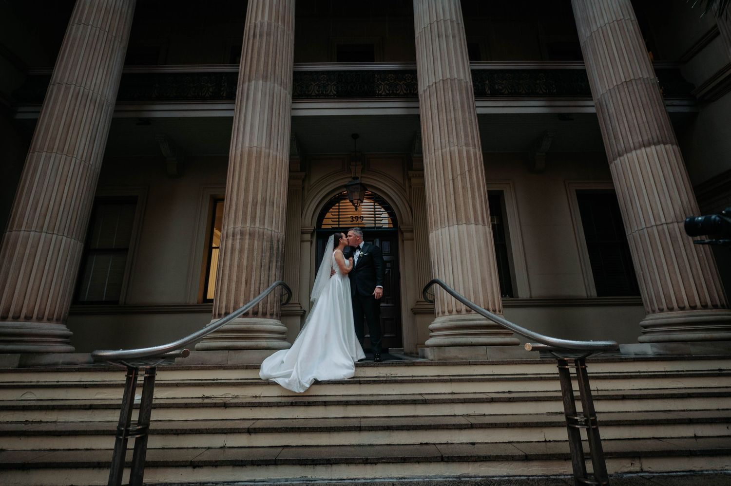 A couple in wedding attire shares a romantic moment between classic stone columns at a grand historic building.
