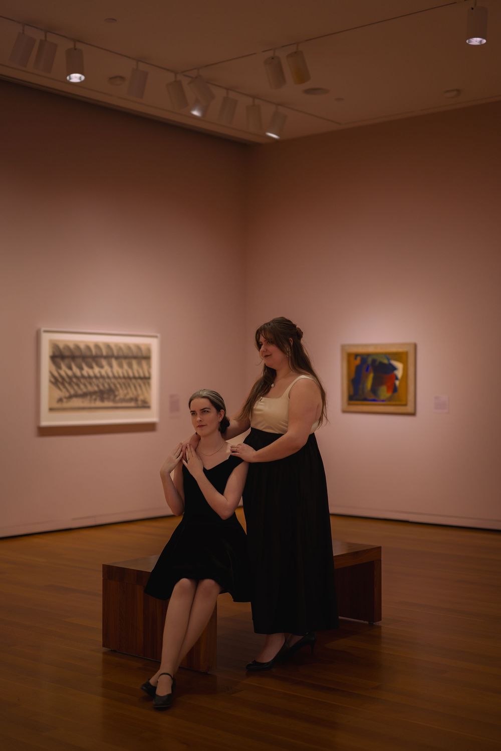 Two women in black dresses pose in an art gallery with paintings and artwork displayed on pink walls.