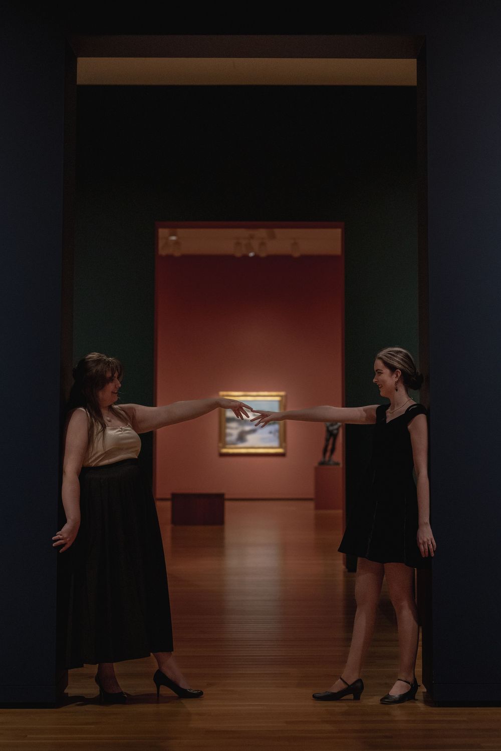 Two people in black dresses gesture towards a small illuminated artwork on a red wall in a dimly lit gallery space.
