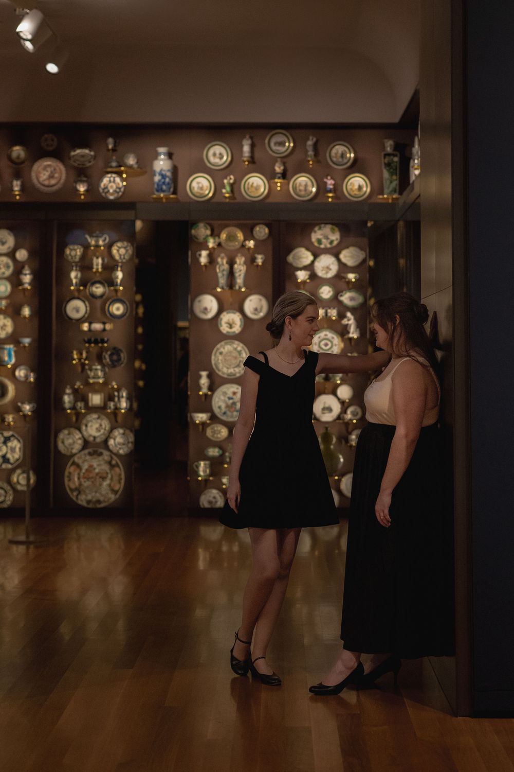 Two women in black dresses stand in front of an illuminated wall display of antique plates and china in an elegant gallery space.