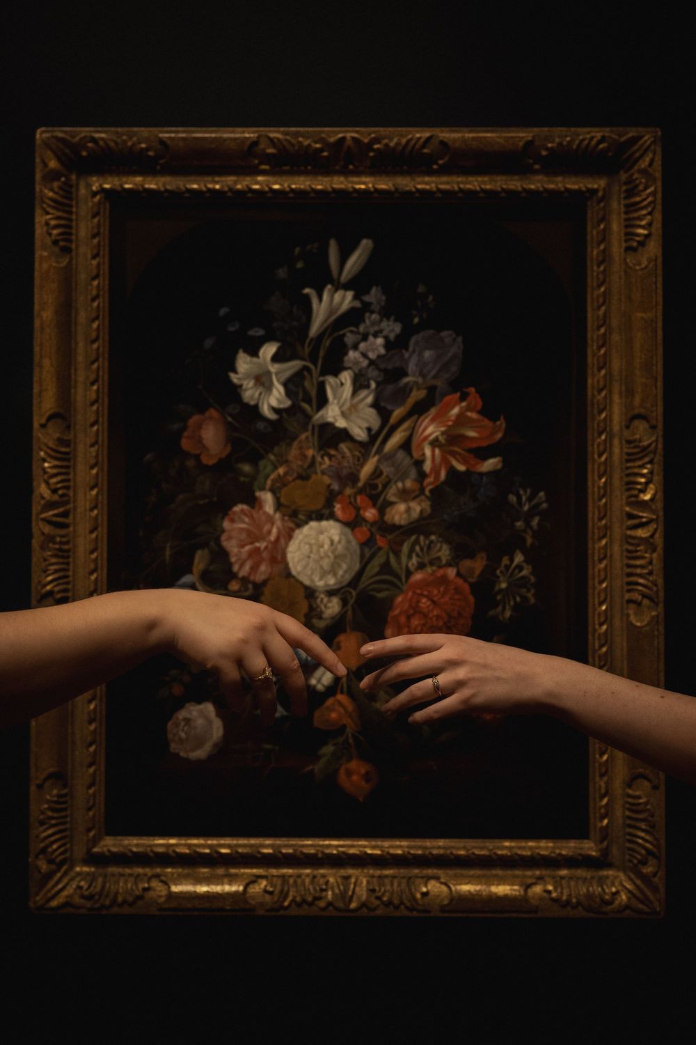Two hands reach toward each other in front of an ornate framed classical still life painting of flowers against a dark background.
