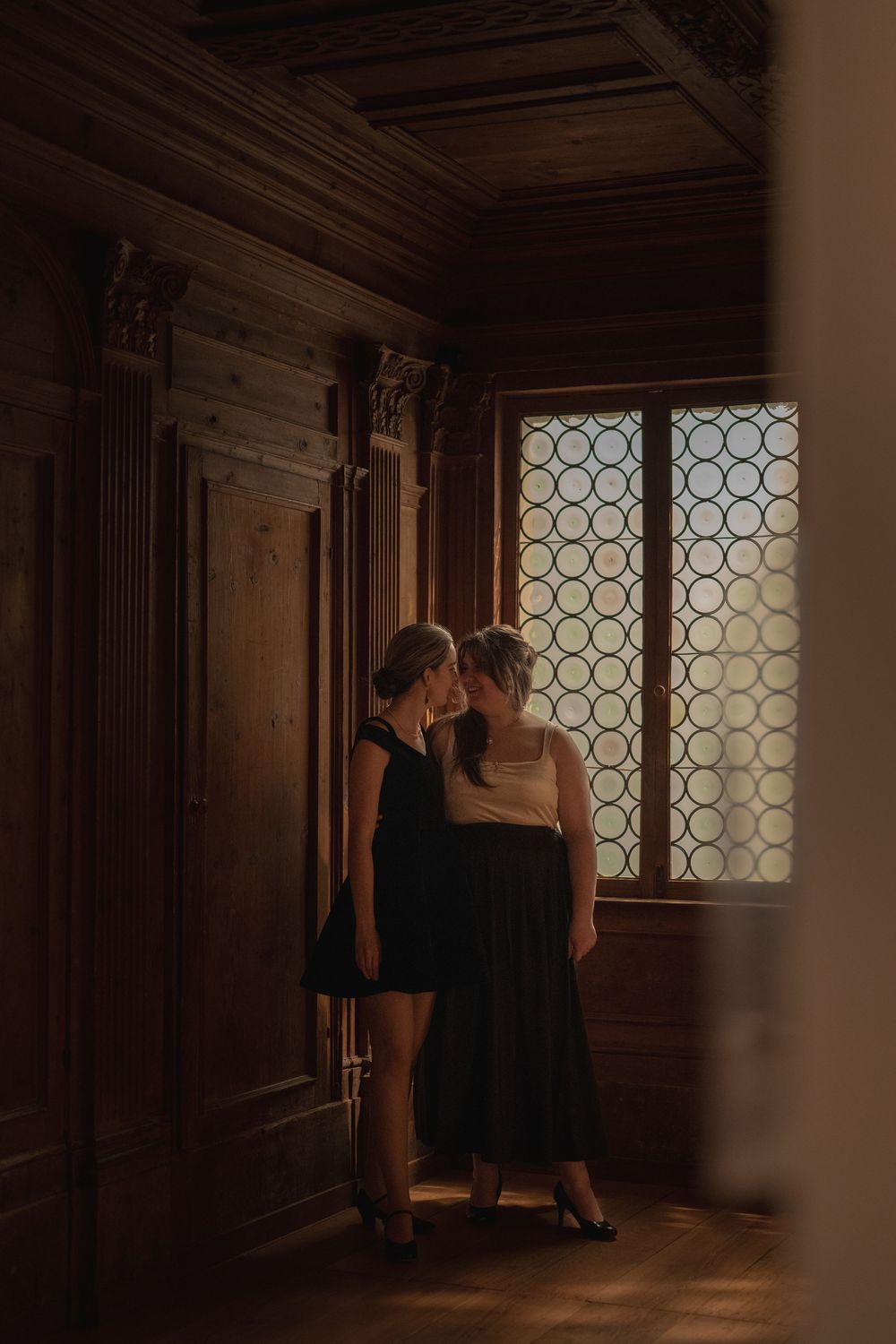 Two figures in black dresses stand intimately near an ornate window in a dark, wood-paneled historic interior.