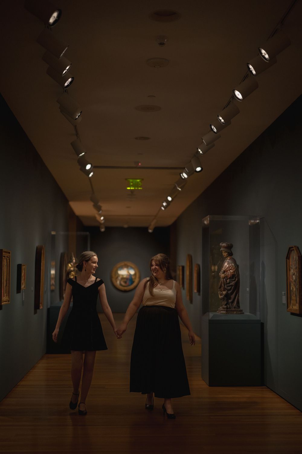Two friends in black dresses holding hands while walking through a dimly lit museum gallery with artwork on display.