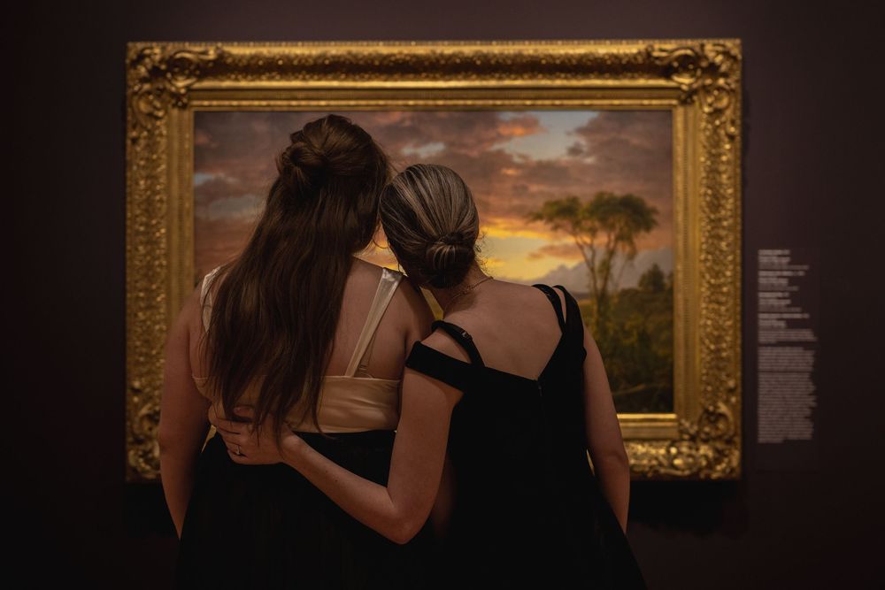 Two museum visitors admire a landscape painting in an ornate gold frame during sunset lighting.
