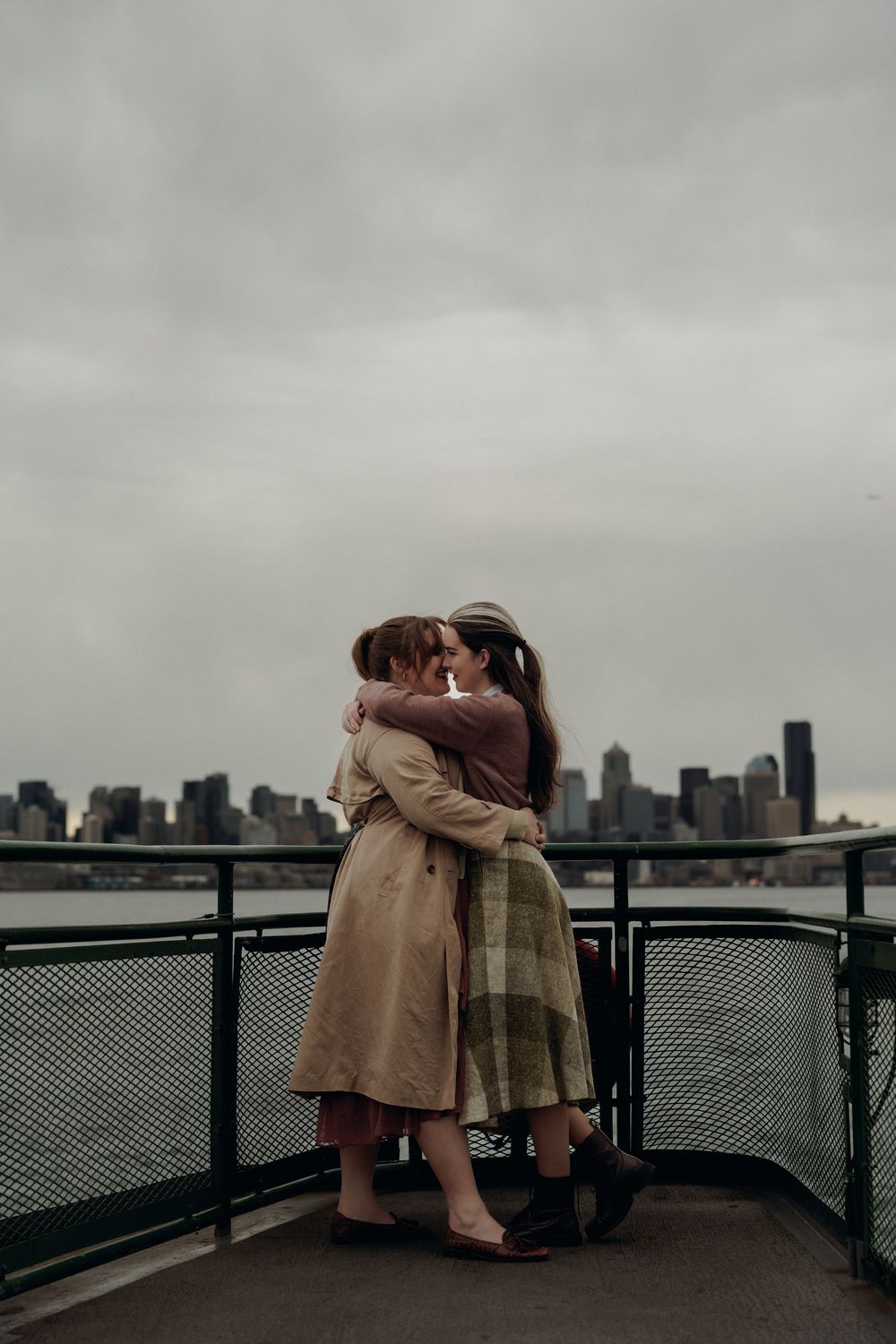 Two people in coats embracing on a viewing deck with a moody city skyline in the background.