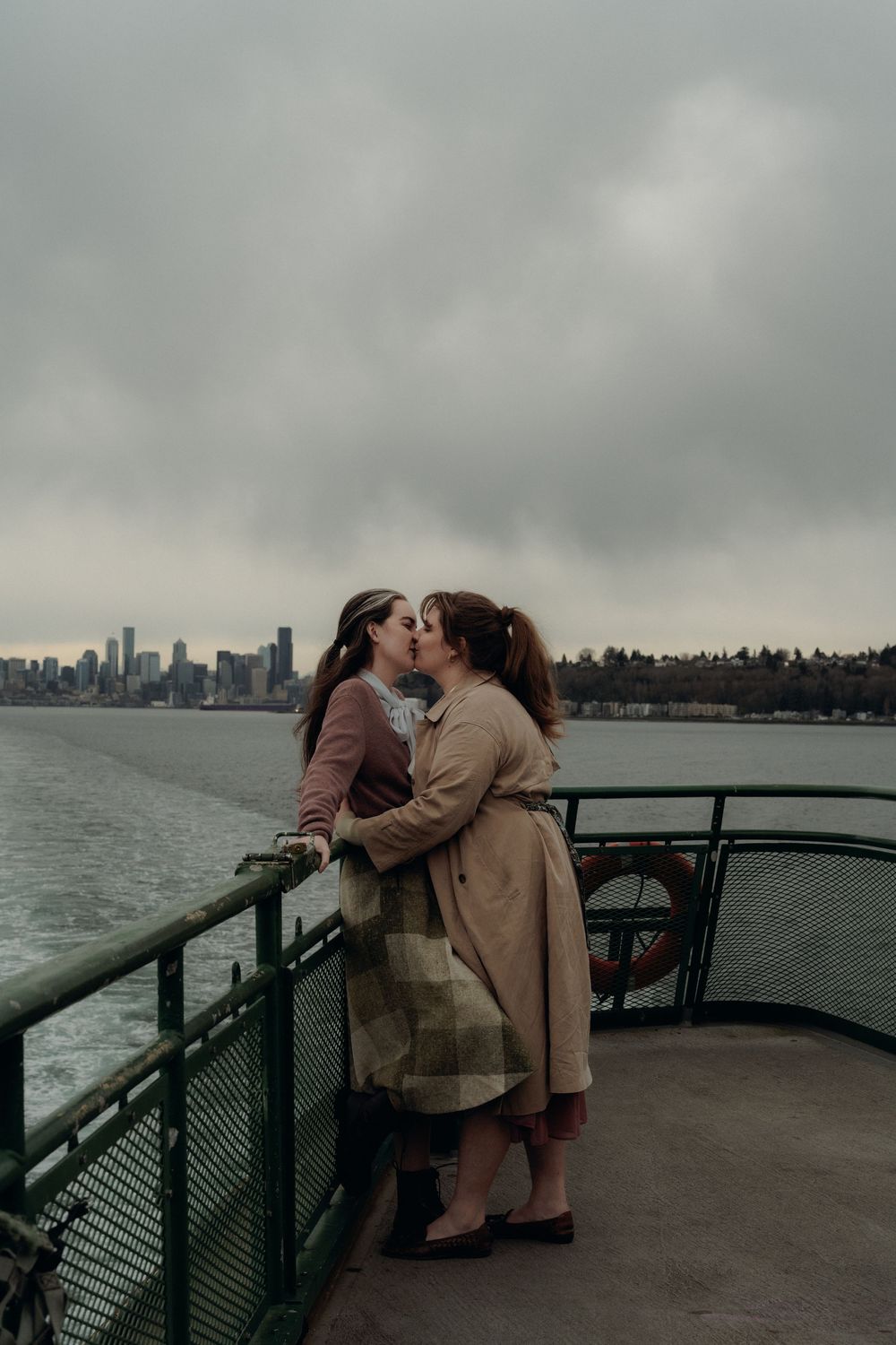 A romantic couple embraces on a ferry deck overlooking the moody Seattle skyline on an overcast day.