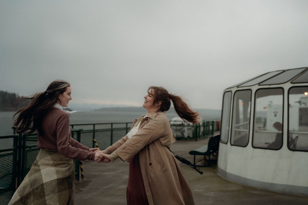 Two figures dance together on a ferry deck in windy conditions wearing long coats on an overcast day near water.