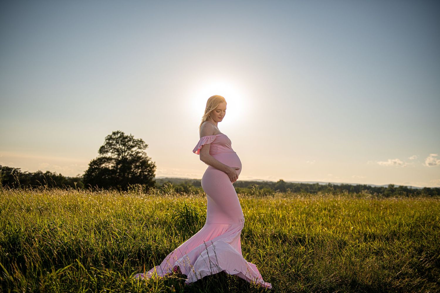 Silhouette of pregnant woman in flowing dress standing in golden sunset field.