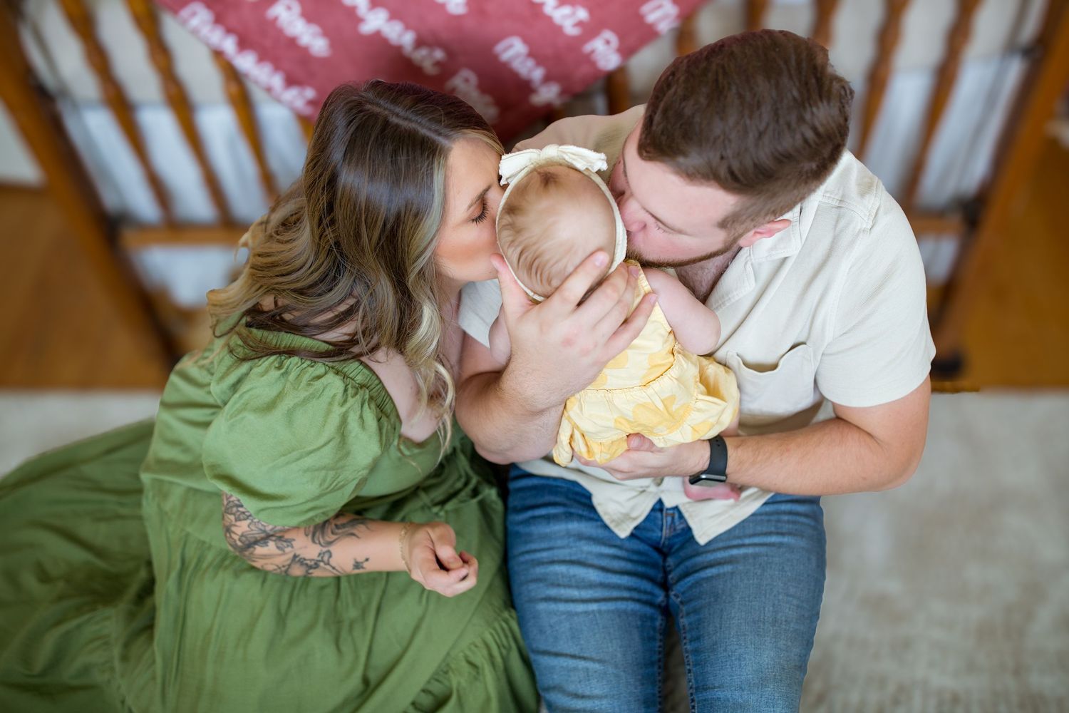 Parents leaning in to kiss their baby girl in a yellow dress during a nursery photo session.