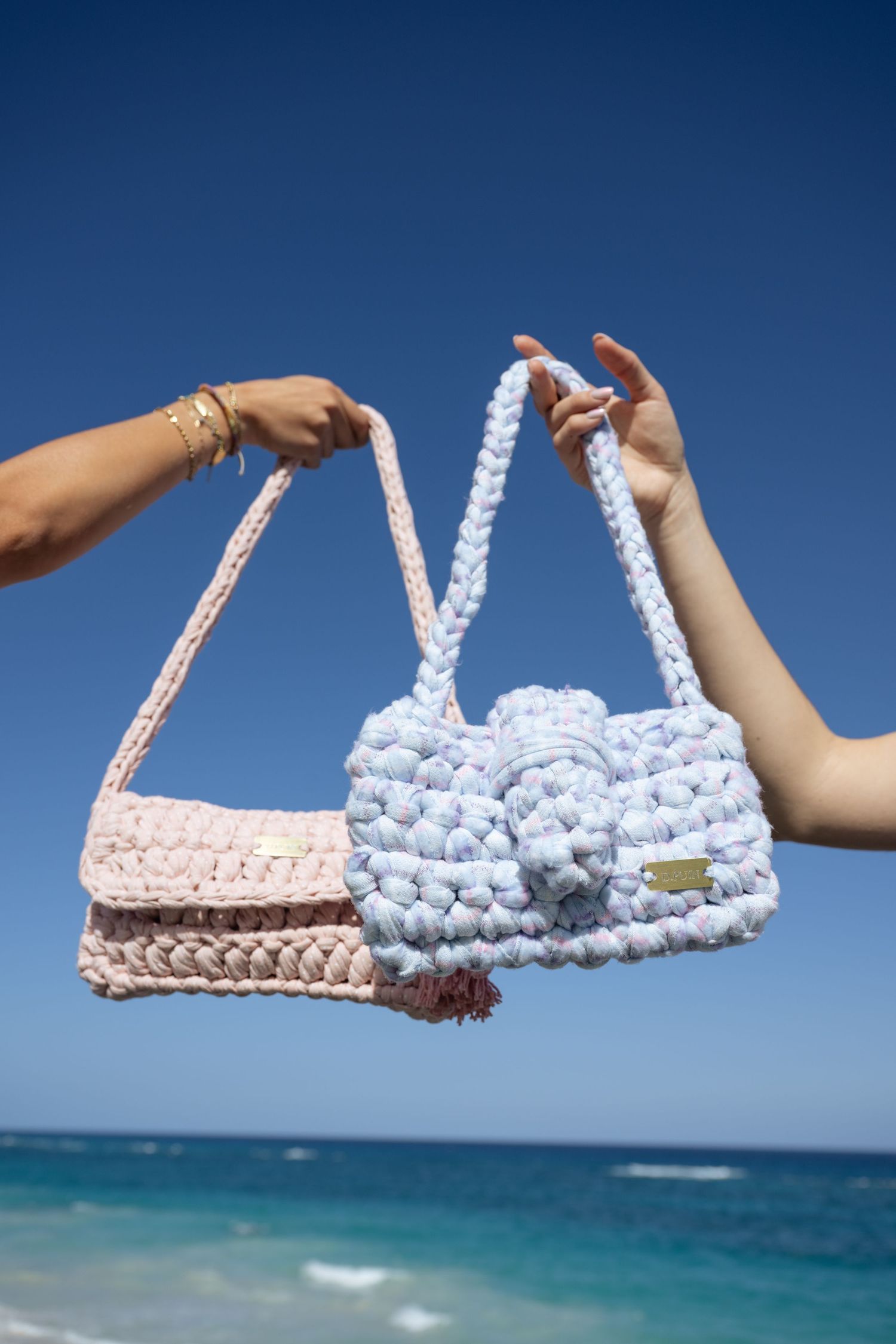 Hands holding up chunky knit handbags against a scenic beach backdrop with blue ocean and sky.