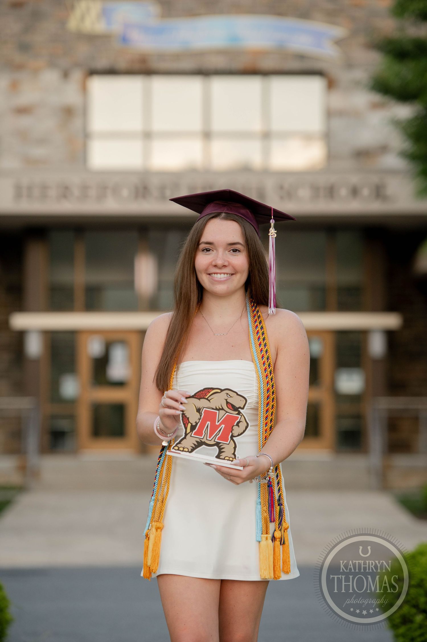 A graduate in a white dress and burgundy cap poses with a university mascot logo and gold honor cords outdoors.