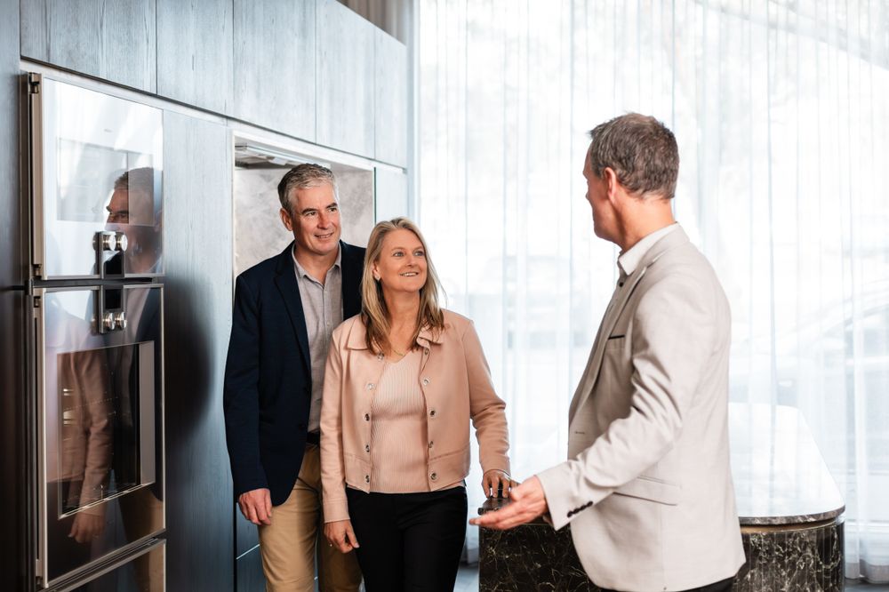 Modern sleek kitchen showroom with wood paneling and two people viewing appliances and fixtures.