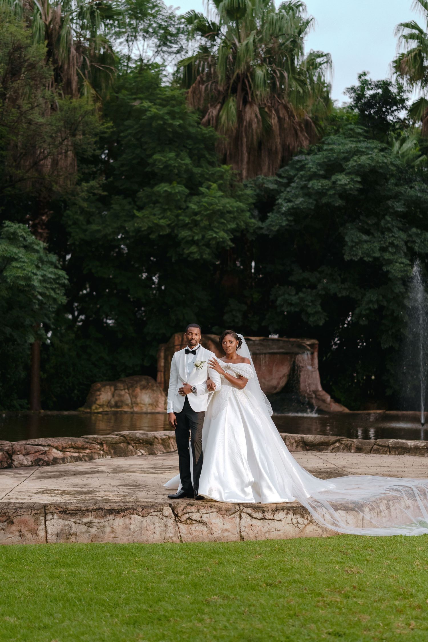 A wedding photo taken outdoors with beautiful greenery, palm trees and stone architecture in the background and a flowing white gown.