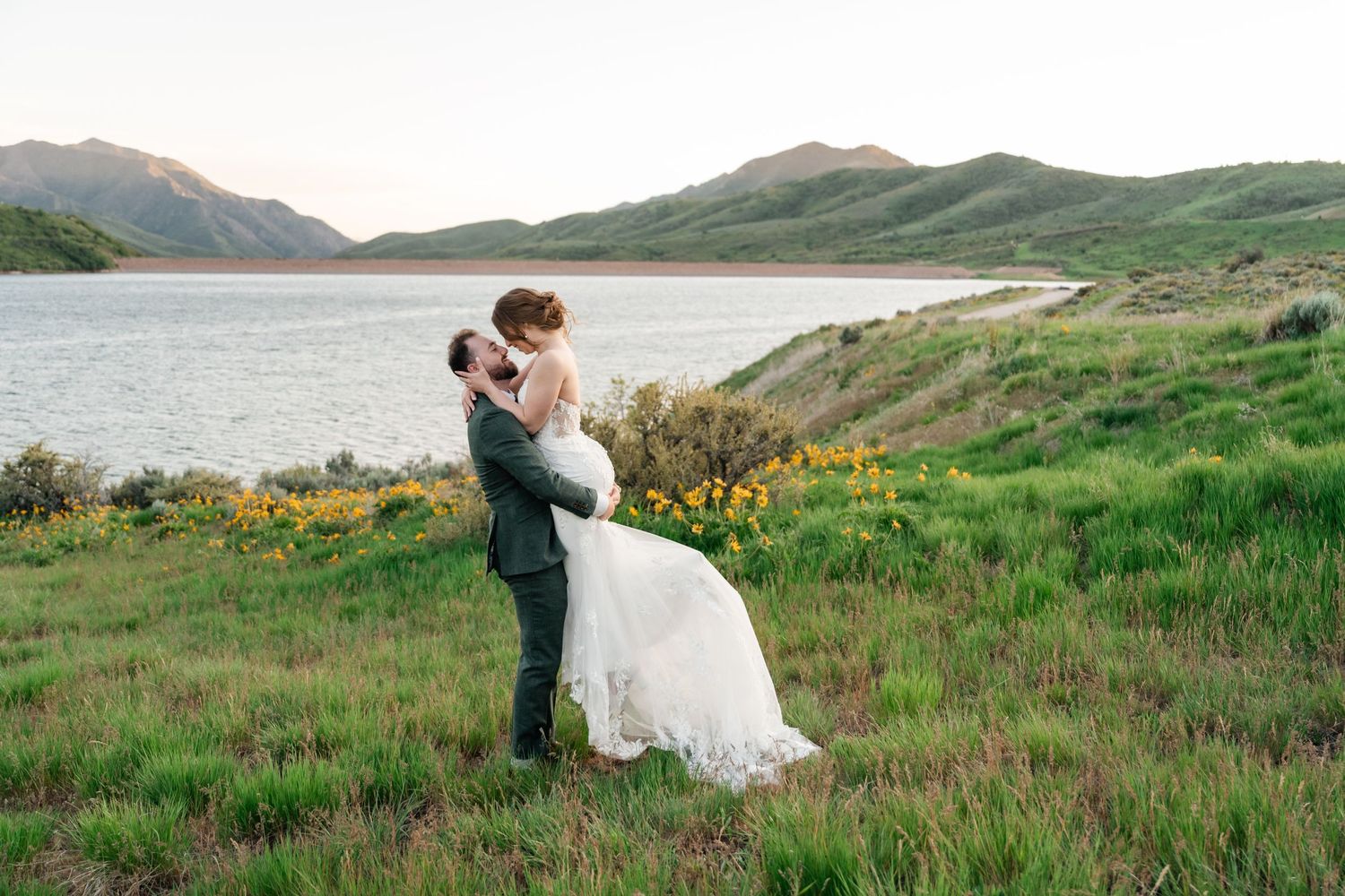 A couple embraces in a romantic moment on grassy hillside overlooking a serene mountain lake with wildflowers blooming nearby.