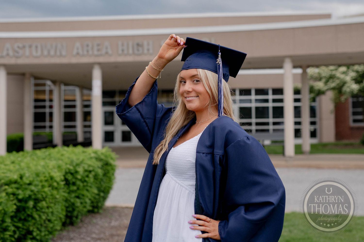 A graduate in blue cap and gown moves their tassel while standing outside a brick building on graduation day.